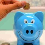 Hand inserting a coin into a blue piggy bank for savings and money management., tags: die zwischen und - pexel
